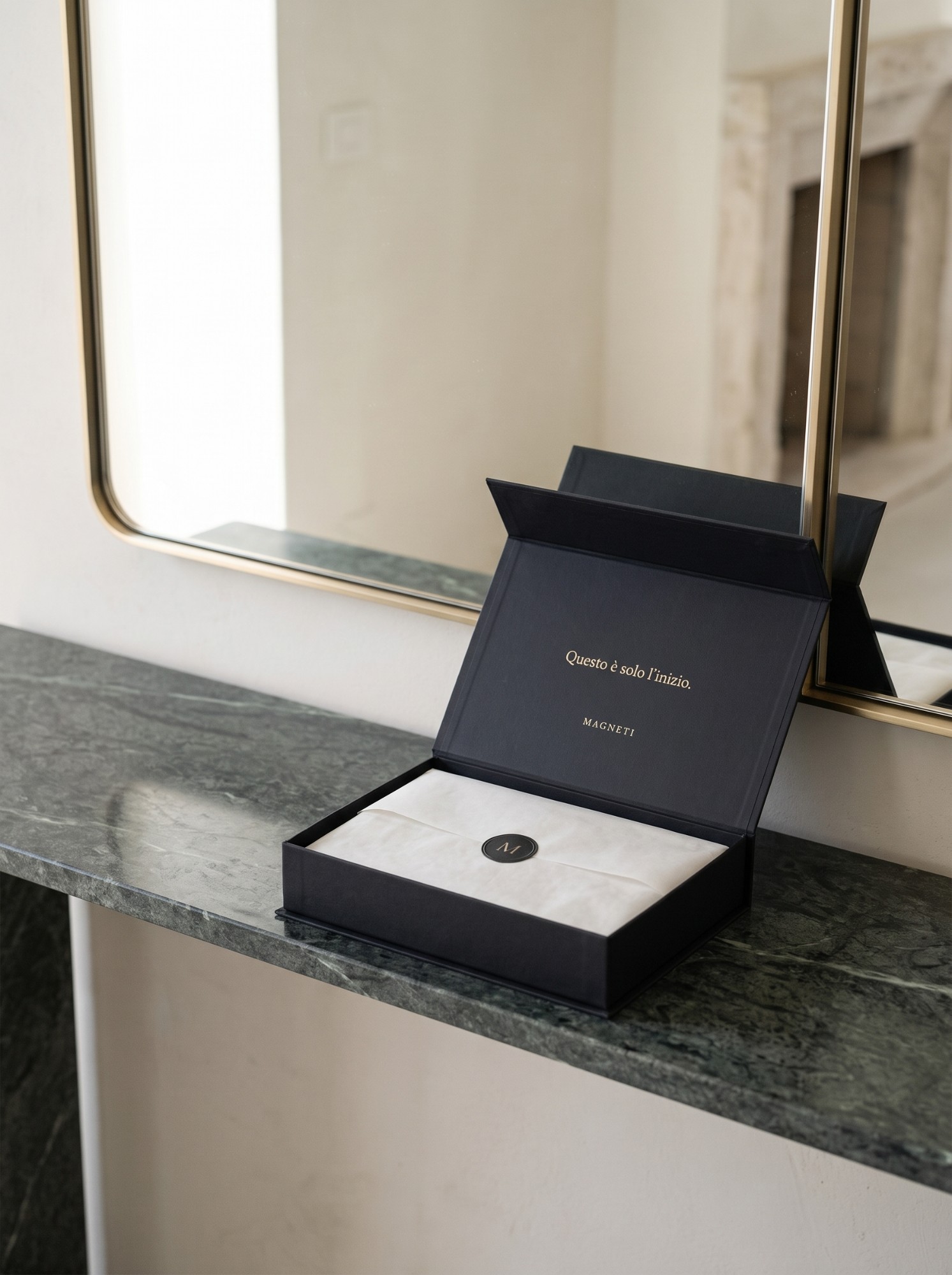 The Magneti® Gift Card box open on a green marble console, in front of a mirror, with the tissue paper and the M seal