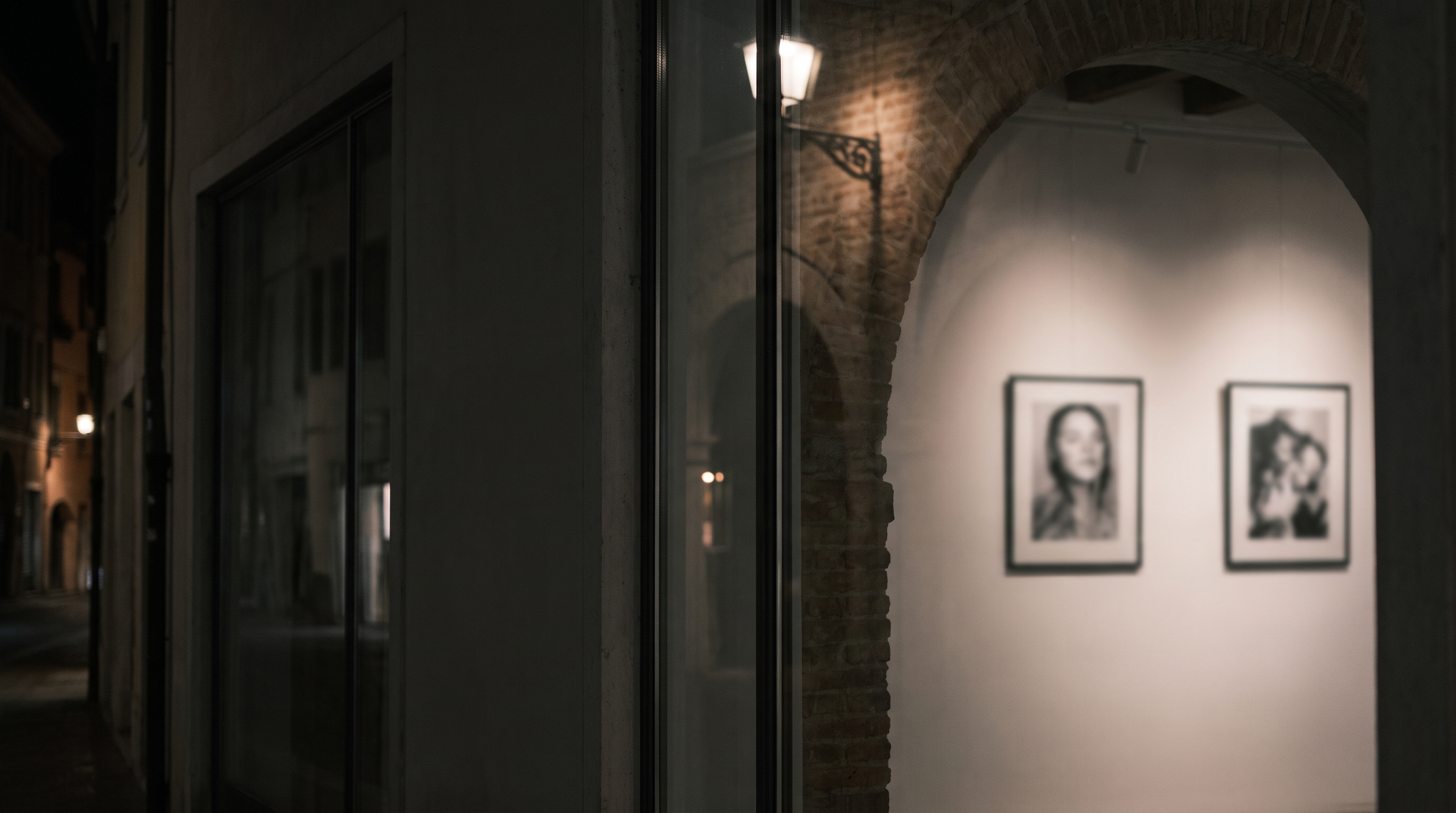 La galleria Magneti® di sera, dalla strada: oltre la vetrina e l’arco di mattoni, due ritratti in bianco e nero sulla parete illuminata