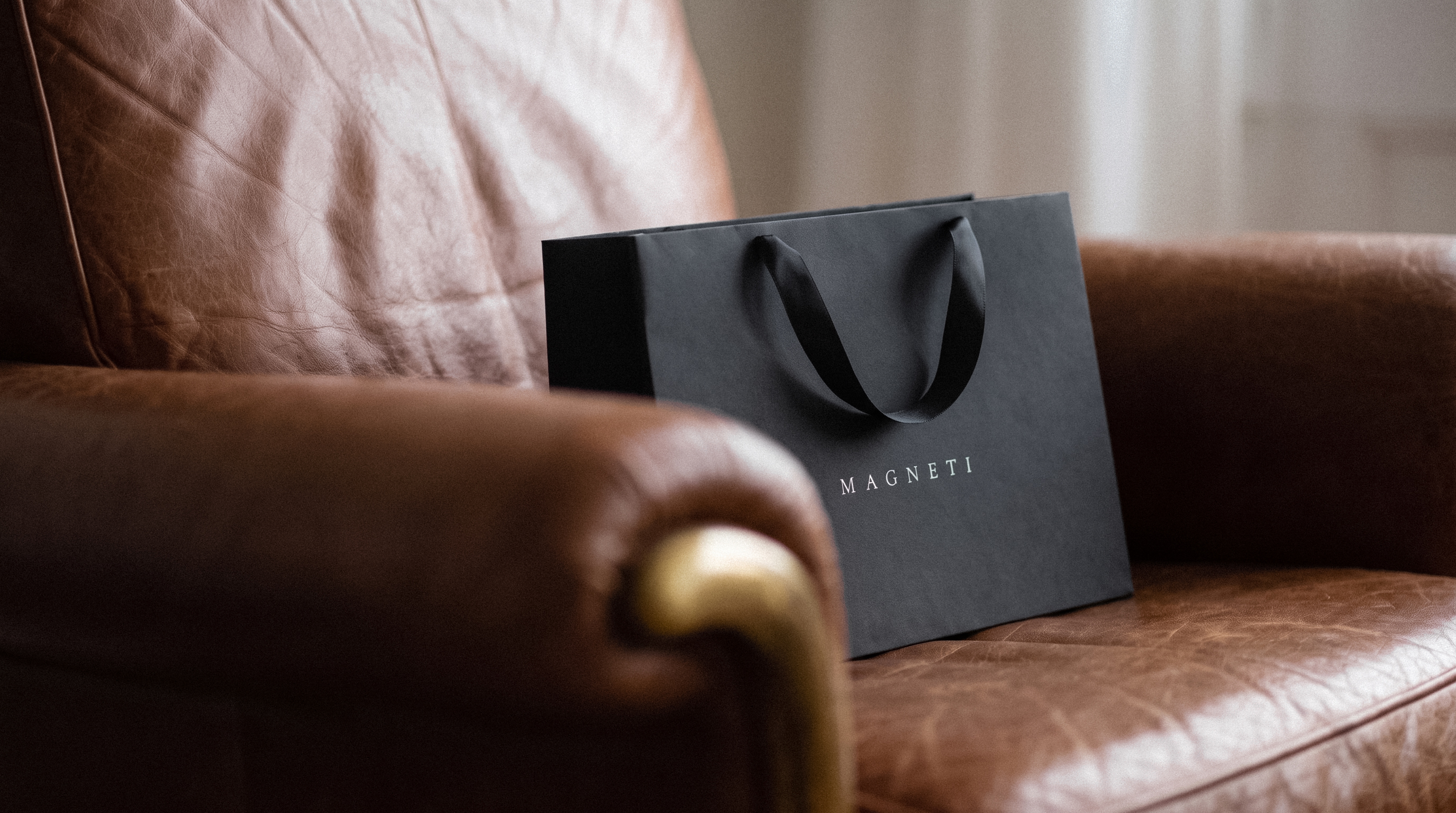The black Magneti® shopping bag with satin handles, resting on a leather armchair